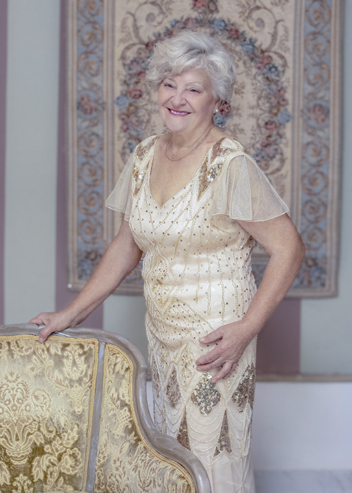 Senior woman in vintage gown standing by an antique chair. Fashion photography