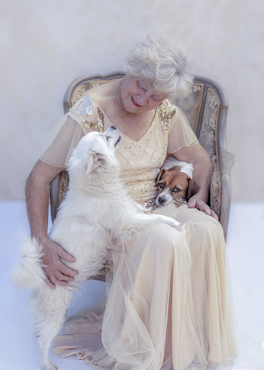 Senior woman in vintage gown sitting in an antique chair with one dog on her lap and the other standing up on her hind legs beside her