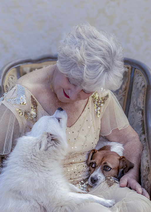 Senior woman in vintage gown sitting in an antique chair with one dog on her lap and the other standing up on her hind legs beside her, close up portrait